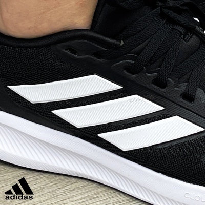 Ténis desportivo Adidas preto com riscas brancas em fundo de chão de madeira