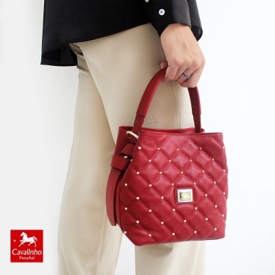 Mala de mão vermelha acolchoada com detalhes dourados, sendo segurada por uma pessoa com calças bege e camisa preta.