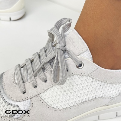 Ténis brancos e cinza com atacadores cinzentos e sola branca, marca GEOX visível no lateral, perna da pessoa calça o sapato.