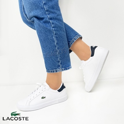 Ténis brancos Lacoste em pele com detalhe verde e preto usados por pessoa com jeans azuis em fundo branco