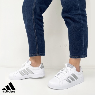 Sapatilhas Adidas brancas com riscas prateadas e jeans escuro num fundo branco