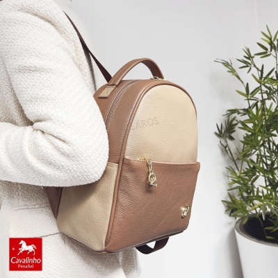 Mochila bege e castanha em pele sintética com textura granulada e zíper dourado, usada com roupa branca texturada ao fundo planta em vaso