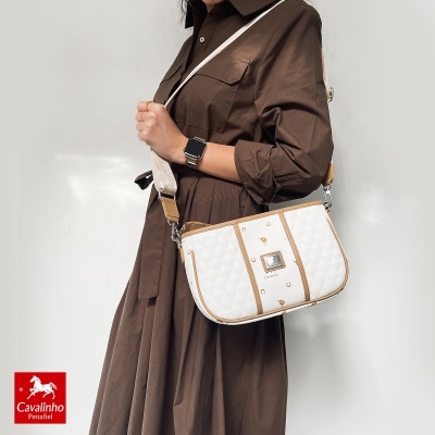Mala de ombro branca e bege Cavalinho Penafiel com padrão de ferraduras sendo usada por modelo com vestido castanho