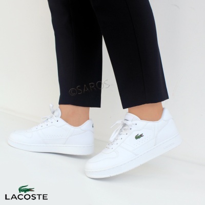 Ténis brancos Lacoste com logótipo verde, usados com calças pretas, fundo branco