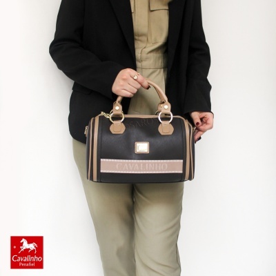 Mala de mão preta e castanha da marca Cavalinho a ser segurada por uma mulher vestida com casaco preto e calças bege