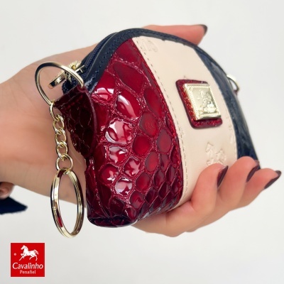 Carteira pequena ou porta-moedas com padrões de pele vermelha, bege e azul, fechada com fecho dourado e argola para chave, na mão.