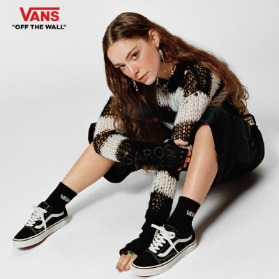 Pessoa com ténis Vans pretos e brancos e meias pretas com logotipo Vans em fundo branco