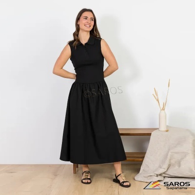 Mulher a vestir vestido preto sem mangas e sandálias pretas, em interior com chão de madeira e decoração minimalista.