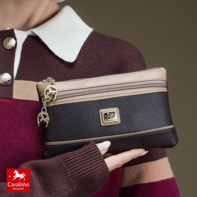 Carteira preta e castanha com fecho dourado em mão de pessoa com camisola castanha, bege e rosa