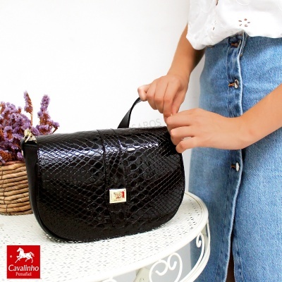 Mala preta estilo crocodilo sobre mesa branca decorativa ao lado de cesto com flores lilases.