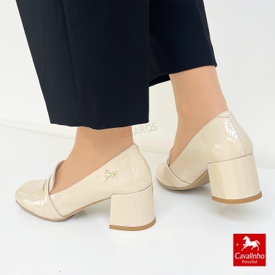 Sapatos de salto médio em verniz bege com detalhe dourado da marca usados com calças pretas.