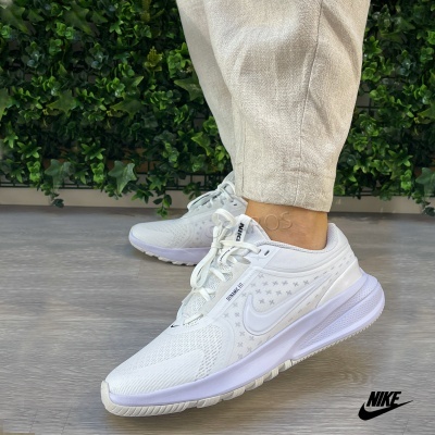 Ténis brancos Nike com malha respirável e sola branca, usados com calças claras num piso de madeira com plantas verdes ao fundo