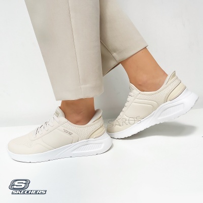 Ténis Skechers bege com sola branca usados por uma pessoa com calças claras num fundo branco.