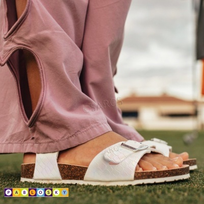 Sandálias brancas com duas tiras e fivela prateada na sola de cortiça, em piso verde ao ar livre