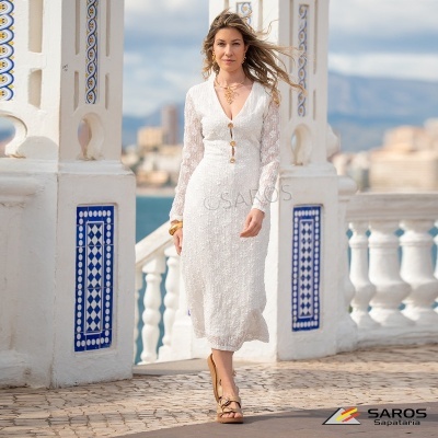 Mulher com vestido branco de malha e sandálias bege em cenário com azulejos azuis e vista de mar