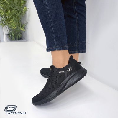 Ténis Skechers pretos em tecido respirável com sola preta, logotipos nas laterais, usados por pessoa com calças de ganga azul escuro