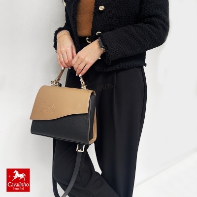 Mala bicolor bege e preta com cavalo em relevo e alças, sendo segurada por mulher vestida com casaco preto e calças pretas.