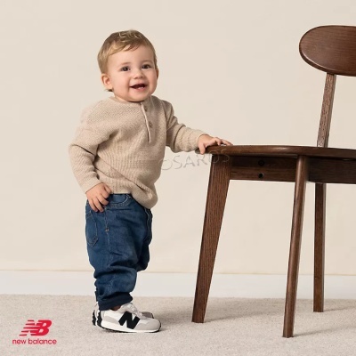 Criança pequena de pé junto a cadeira de madeira, vestindo camisola bege, calças de ganga e ténis New Balance.