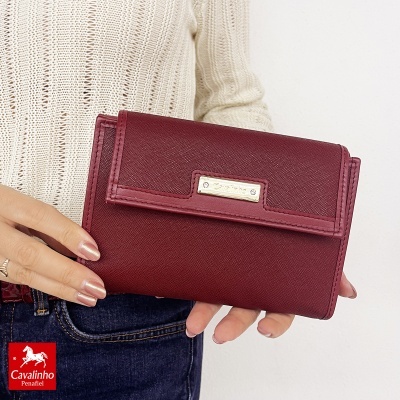 Carteira feminina vinho com placa metálica Cavalinho, segurada por pessoa com camisola branca e jeans.