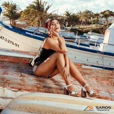 Mulher sentada na praia junto a barcos de pesca, usando top preto, calções cinzentos, sandálias brancas e lenço vermelho na cabeça