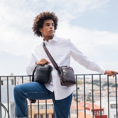 Homem com mala de couro preta ou castanha, camisa branca, calças de ganga, cenário urbano ao fundo