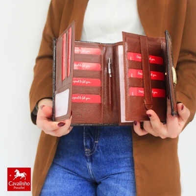 Carteira em pele castanha aberta com vários compartimentos para cartões vermelhos e texto branco segurada por pessoa com casaco castanho e jeans azuis