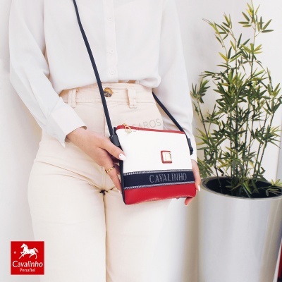 Mala de ombro pequena Cavalinho branco, vermelho e preto usada por pessoa com roupa clara ao lado de planta