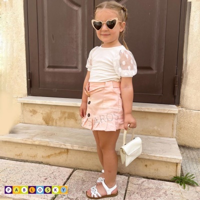 Criança com saia rosa, camisa branca, óculos de sol de coração, sandálias e bolsa branca