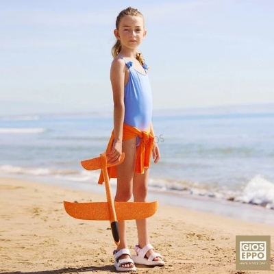 Menina na praia com fato de banho azul claro e sandálias brancas segurando brinquedo avião laranja