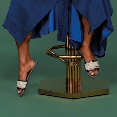 Pés femininos com sandálias de tiras largas brancas e cinza, saia azul, sentada numa cadeira dourada sobre fundo verde.