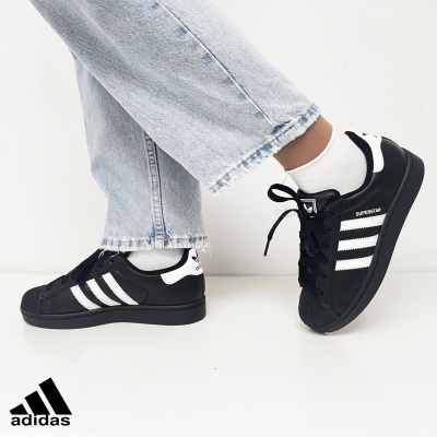 Ténis Adidas Superstar pretos com riscas brancas e sola preta usados com meias brancas e jeans claros