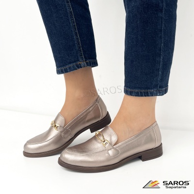 Sapatos tipo mocassim femininos dourados claros com corrente dourada e sola castanha, usados com calças de ganga azul escuro.