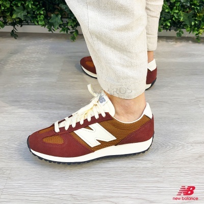 Ténis New Balance castanhos e brancos com sola preta usados por pessoa com calças claras sobre chão de madeira clara perto de plantas.