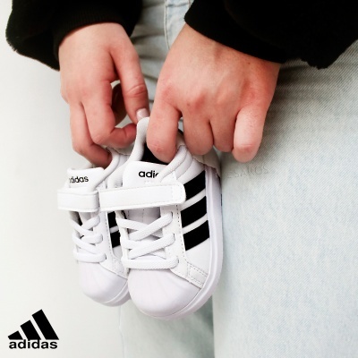 Sapatilhas infantis Adidas brancas com riscas pretas e fecho em velcro nas mãos de uma pessoa.