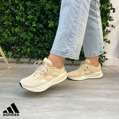 Ténis Adidas bege com três riscas creme usados com jeans claros diante de parede branca e plantas verdes