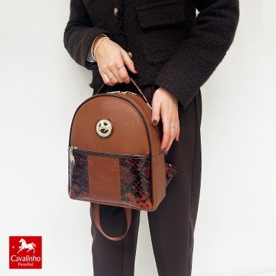 Mochila castanha com detalhes em pele de cobra e fecho dourado segurada por pessoa com casaco castanho escuro