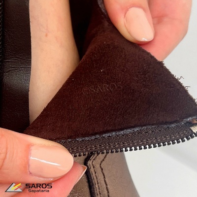 Parte interna de calçado castanho com fecho e selo SAROS, mãos segurando o produto