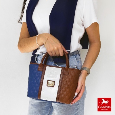 Mala de mão Cavalinho azul, branco e castanho com padrão matelassé e logótipo dourado, segurada por mulher