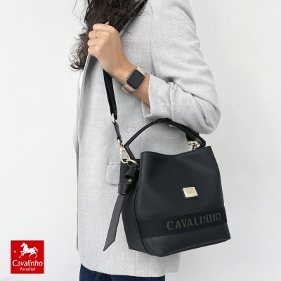 Mala preta em pele com logótipo Cavallinho e alças, usada no ombro de mulher com casaco cinza.