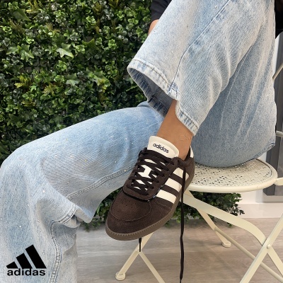 Ténis castanho escuro Adidas com riscas brancas usados com calças de ganga azul clara sentada numa cadeira branca junto a parede com plantas verdes