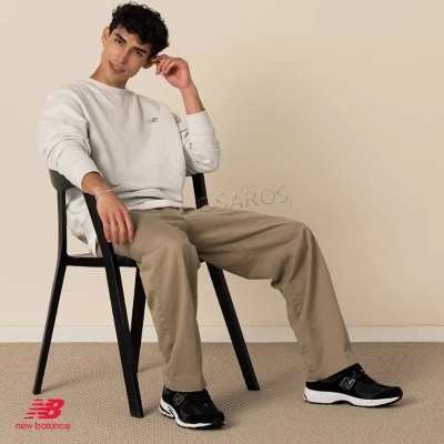 Homem com roupa casual cinza e bege sentado em cadeira preta e sapatilhas negras New Balance.