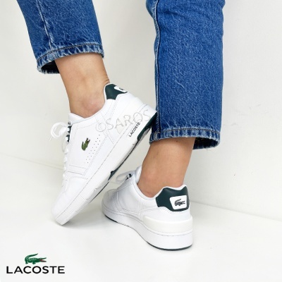 Sapatilhas Lacoste brancas com detalhes verdes e sola branca, usadas com calças de ganga azuis
