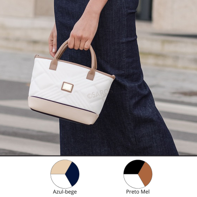 Mala de mão pequena bege e branca com padrão geométrico sendo segurada por mão feminina junto a calças de ganga