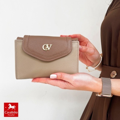 Carteira de pele castanho claro e escuro com fecho e logo dourado, mostrada por mulher com vestido castanho