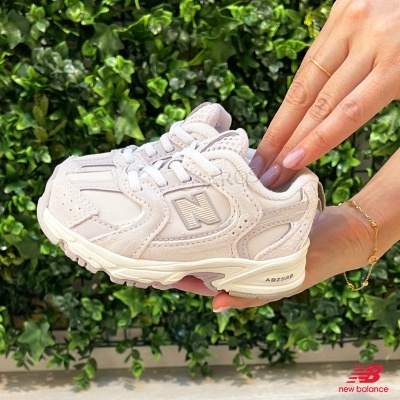 Ténis New Balance bege para criança segurados ao lado de uma parede verde
