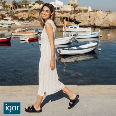 Mulher com vestido branco de malha a caminhar junto a porto com barcos e mar