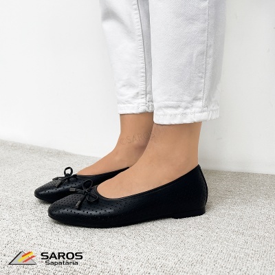 Sapatilhas pretas perfuradas com laço usadas com calças brancas, sobre tapete cinza