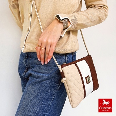 Mala de ombro bicolor Cavalinho Penafiel com alça fina em ombro de pessoa vestindo cardigan bege e jeans azuis
