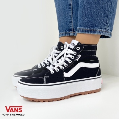 Ténis altos Vans pretos com sola branca e risca lateral branca usados com calças de ganga azuis.