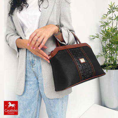 Mala preta com detalhes castanhos e padrão de cavalos numa faixa central, segurada por mulher vestindo casaco cinza e jeans claros.